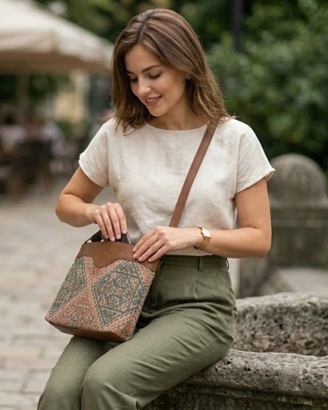 Woman sitting outdoors with a patterned bag, wearing a white shirt and green pants.