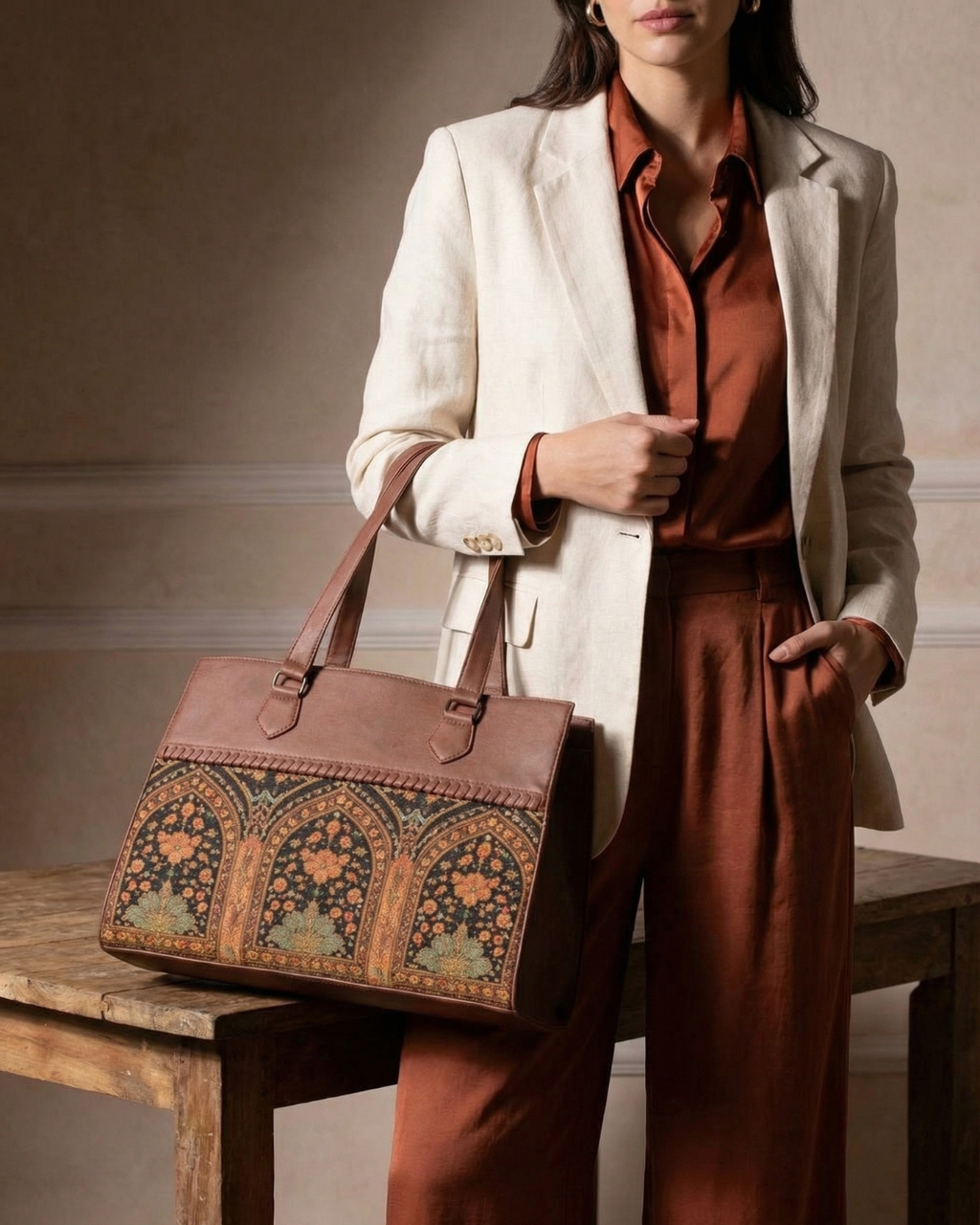 Woman holding a brown leather bag with intricate patterns, wearing a beige blazer and rust-colored pants.